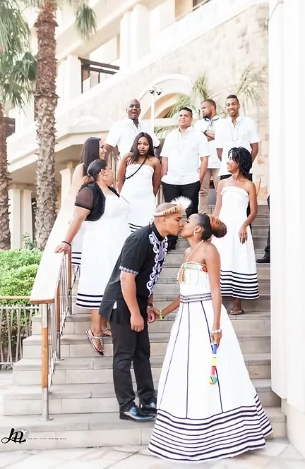 Couple kissing on white steps, the bride in a white dress with black striped bands, bridal party behind.