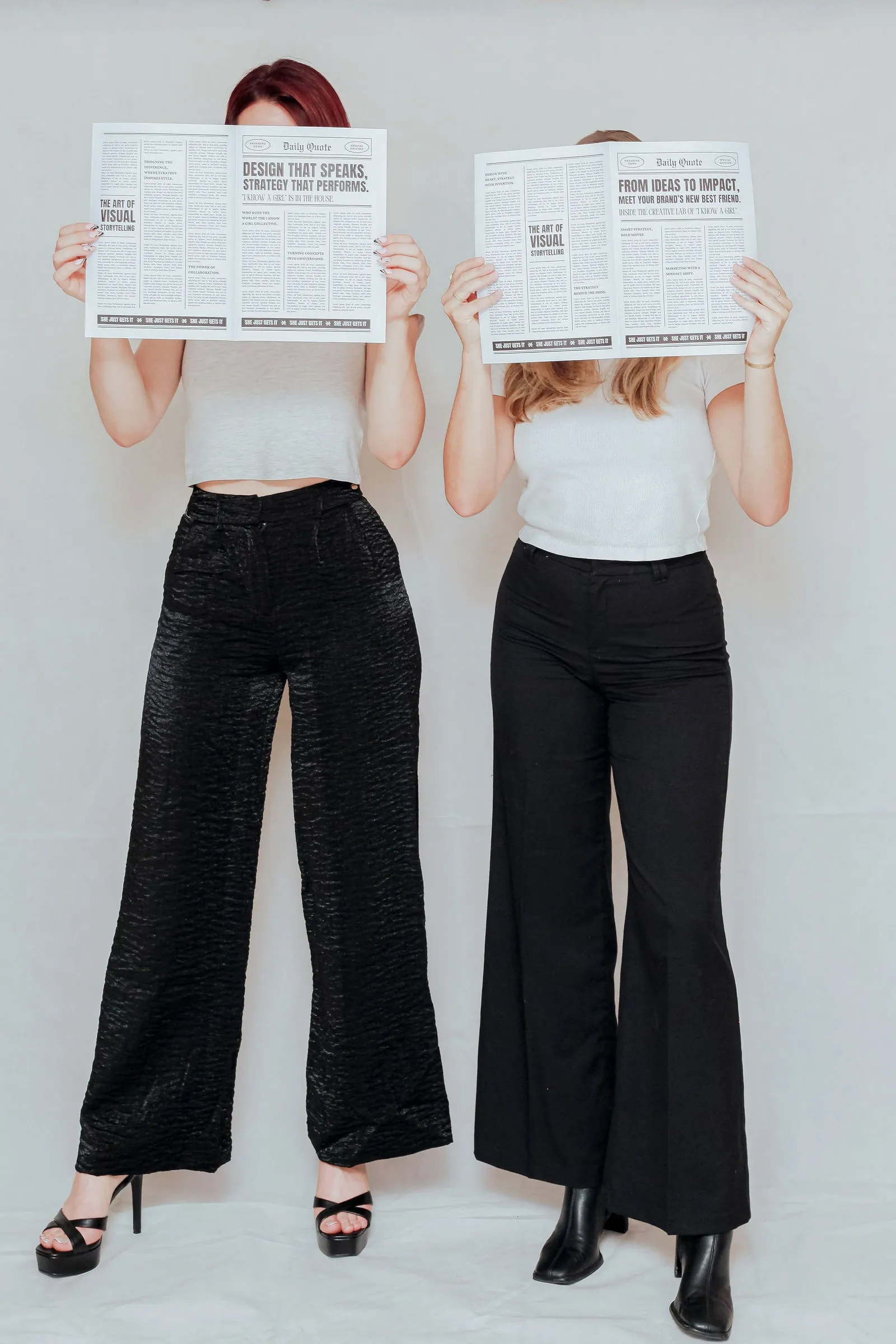 Two women in white tops and black wide-leg trousers standing side by side, faces hidden behind open mock newspapers.