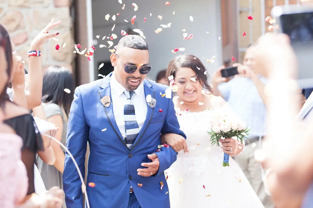 Bride and groom walking out through a shower of rose petals, the groom grinning in sunglasses.