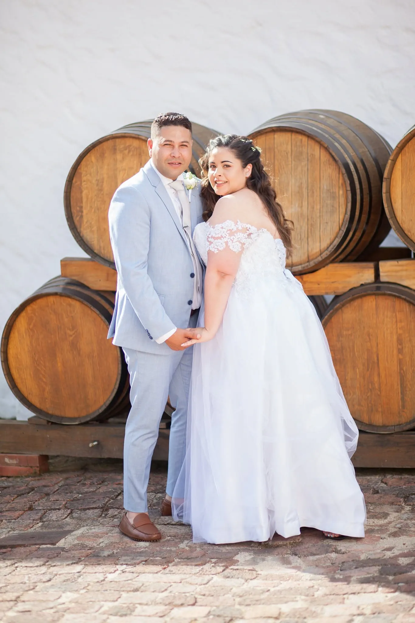Bride and groom holding hands in front of stacked wine barrels.