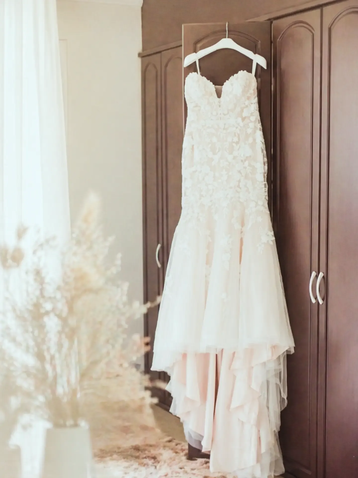 Lace wedding dress on a white hanger against a dark wooden wardrobe door, beside a bright window.