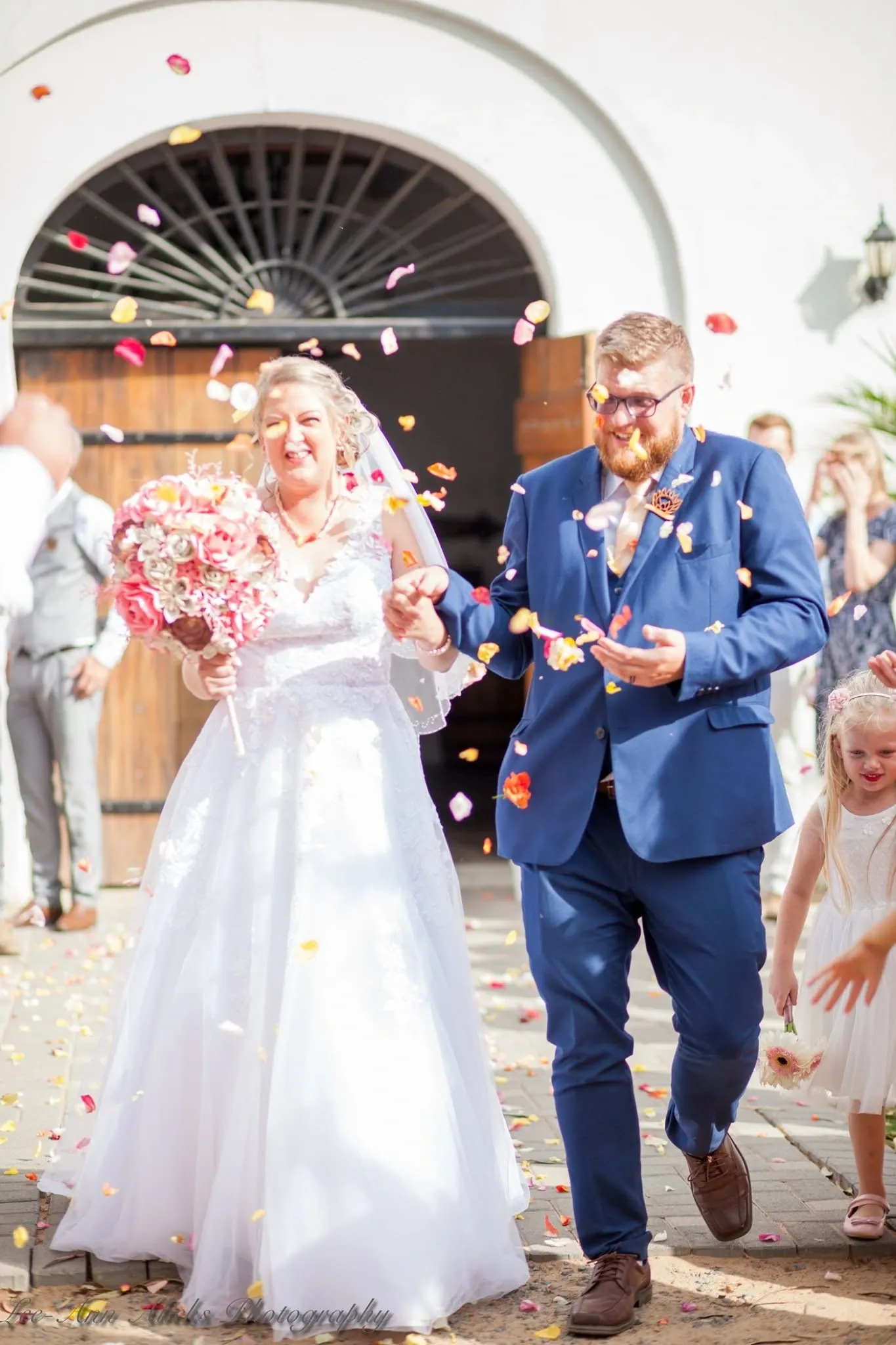 Bride and groom walking out of an arched doorway through a shower of rose petals.