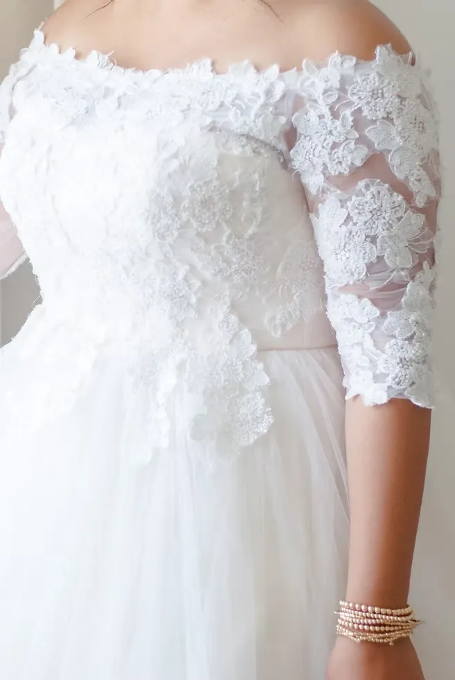 Close-up of an off-the-shoulder bridal gown with lace half-sleeves and a stack of beaded bracelets.